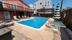 A swimming pool with lounge chairs and a table, surrounded by a wooden deck and a brick patio.