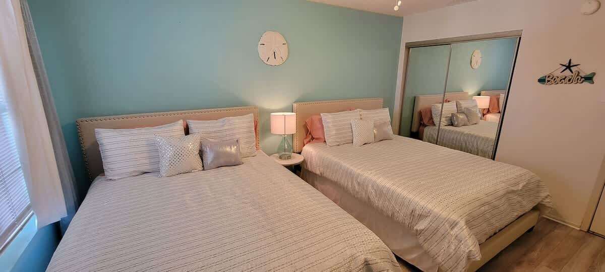 A bedroom with two beds, a bedside table with a lamp, a clock on the wall, and a mirror.