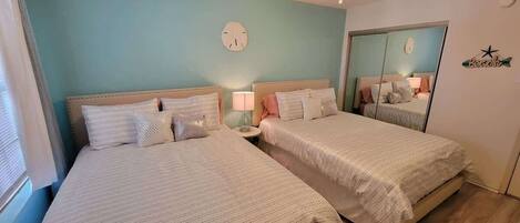 A bedroom with two beds, a bedside table with a lamp, a clock on the wall, and a mirror.