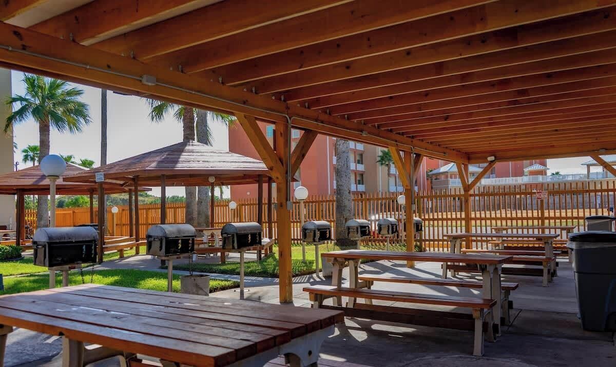 An outdoor patio area with wooden benches and picnic tables, surrounded by a wooden fence and featuring barbecue grills.