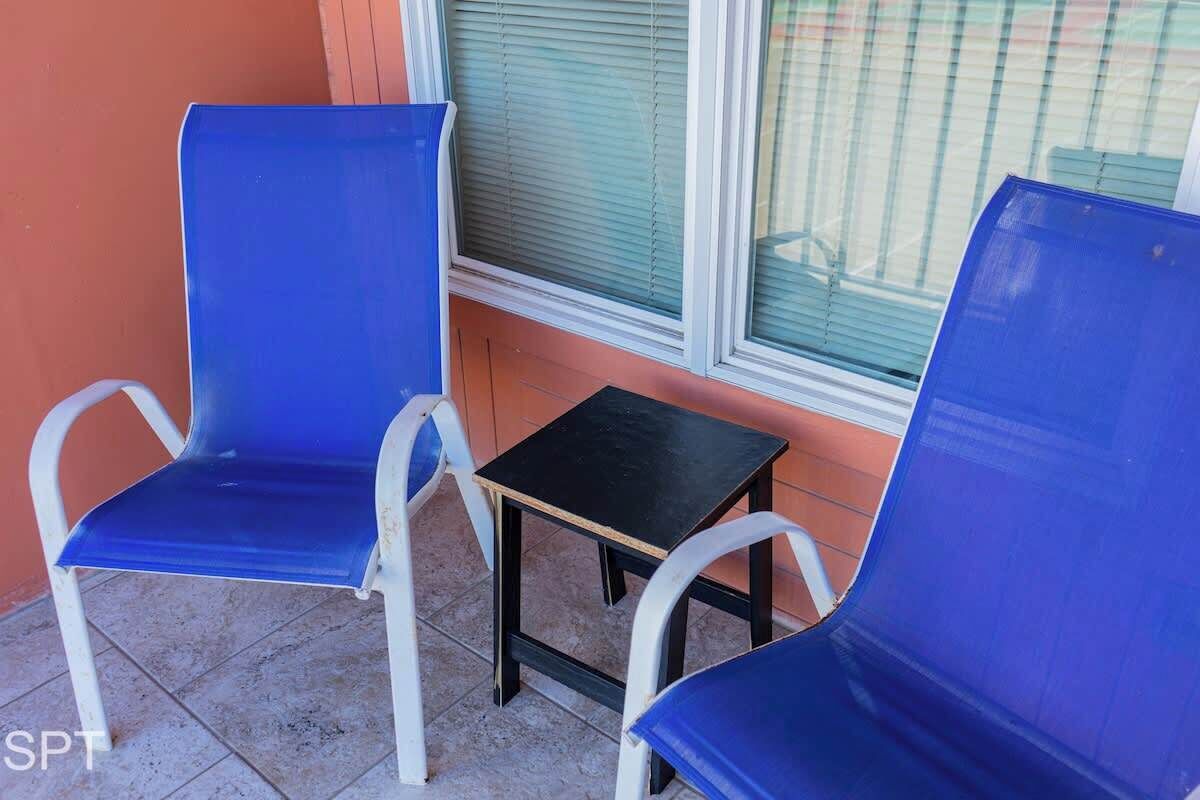 To blå stole og et lille, sort bord på en fliseterrasse med et vindue i baggrunden.