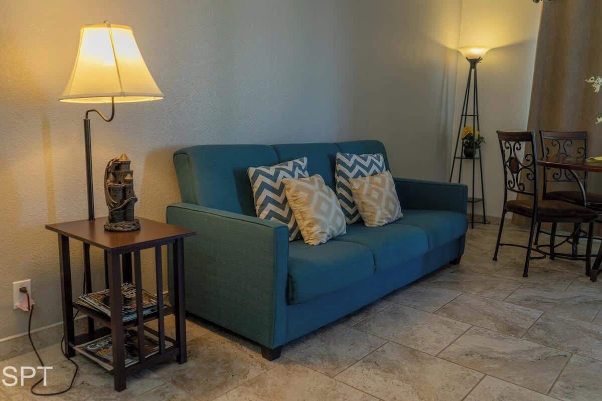 Una sala de estar con un sofá color turquesa, cojines estampados, una mesita con lámpara y una zona de comedor con sillas.