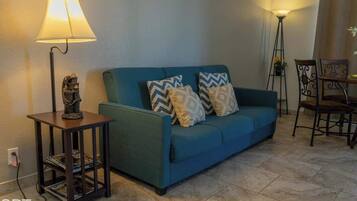 Una sala de estar con un sofá color turquesa, cojines estampados, una mesita con lámpara y una zona de comedor con sillas.