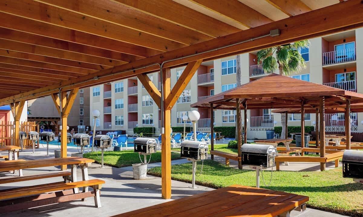 An outdoor patio area with wooden benches and barbecue grills, surrounded by a multi-story building with balconies and a pool.