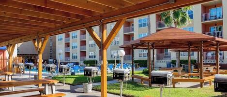 An outdoor patio area with wooden benches and barbecue grills, surrounded by a multi-story building with balconies and a pool.
