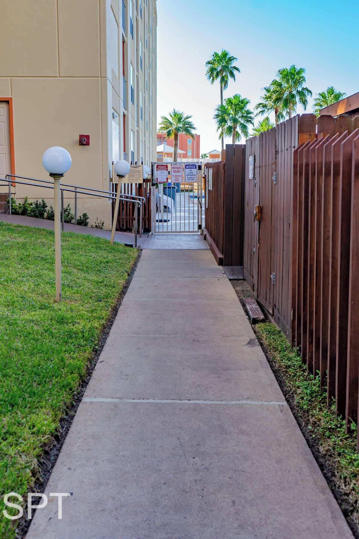 A pathway leading to a gated entrance with a wooden fence, a grassy area, and a building with a beige facade.