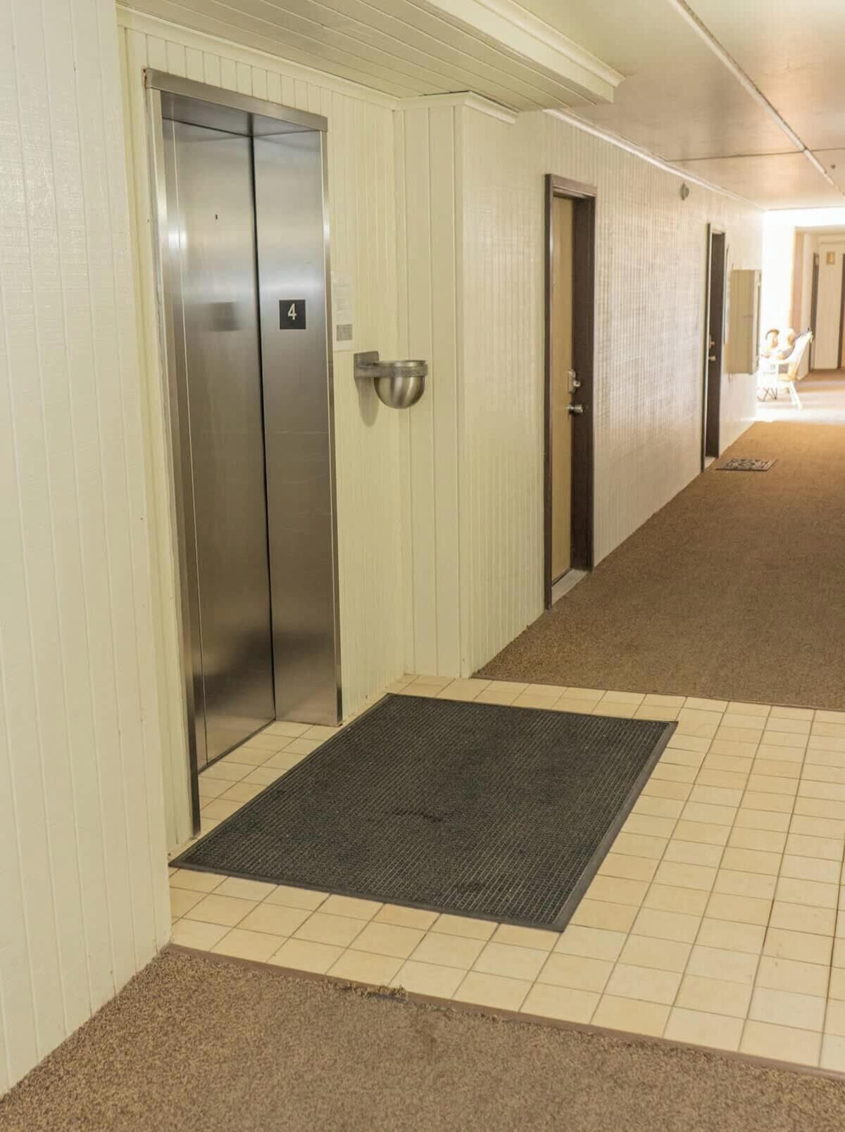 A hotel corridor with a carpeted floor, a metal elevator, and a door with a number plate.