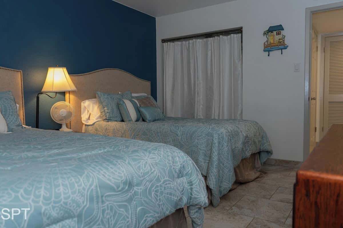 A bedroom with two beds, a bedside table with a lamp, a wall-mounted fan, and a window with curtains.