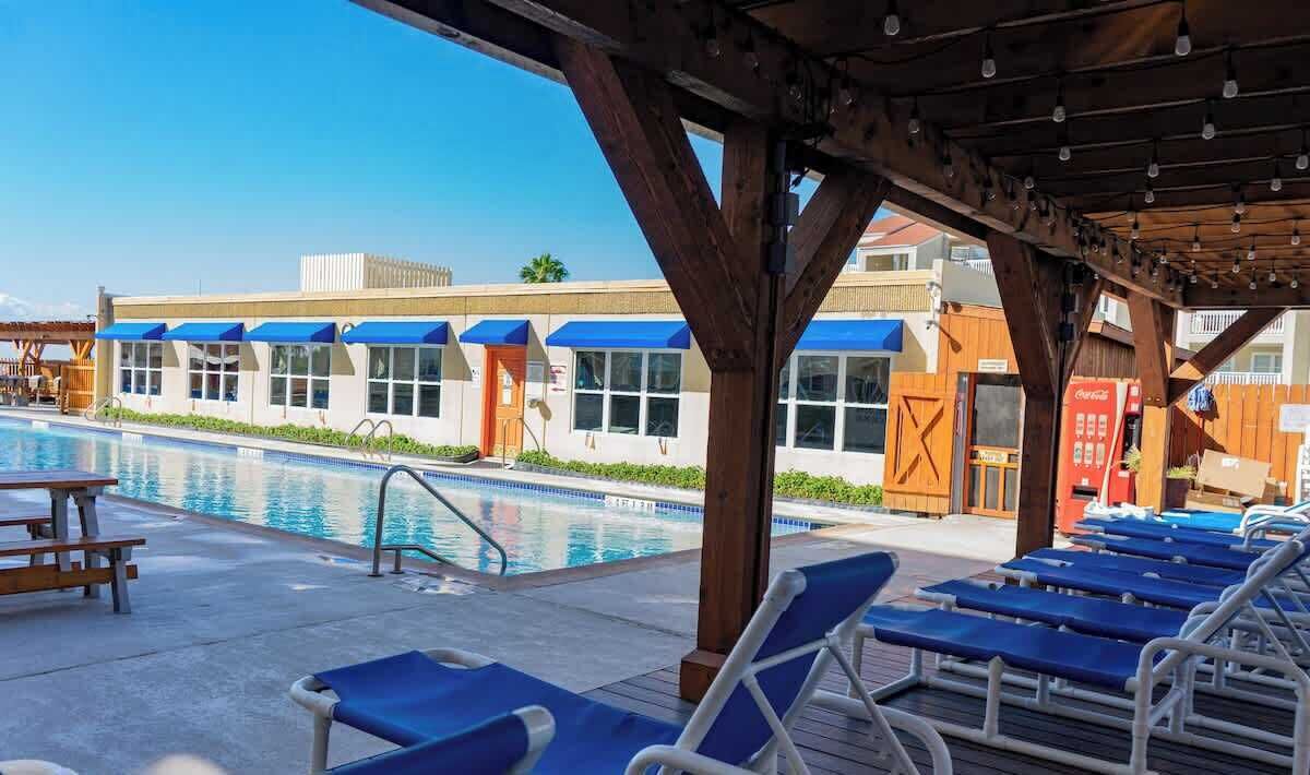Zona de piscina con sillones de descanso, un espacio de estar cubierto y un edificio con toldos azules.
