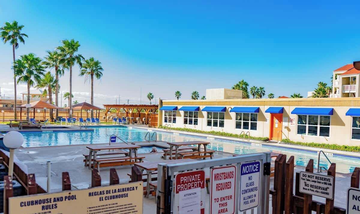 Un área de piscina con sillones de descanso, palmeras y un edificio con toldos azules.