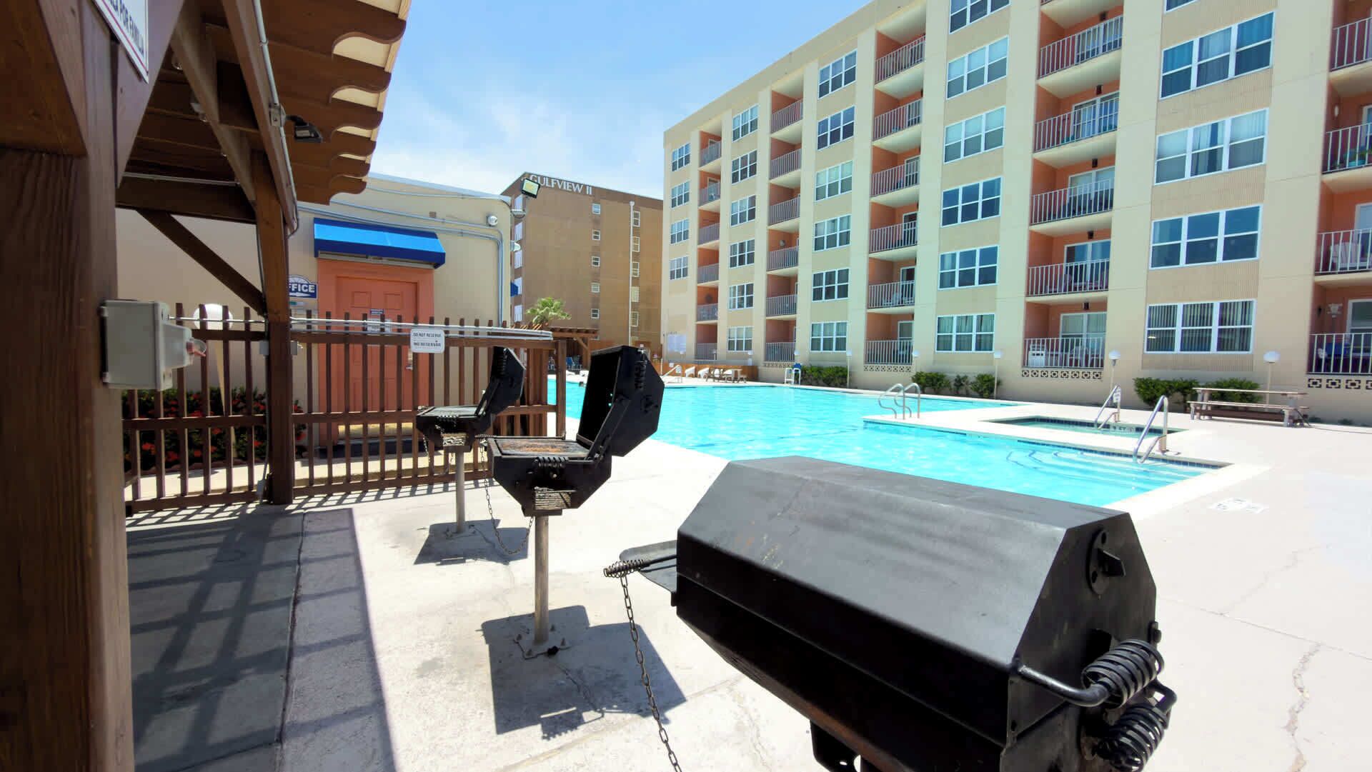 Zona junto a la piscina con una parrilla, mobiliario exterior y un edificio de varios pisos al fondo.