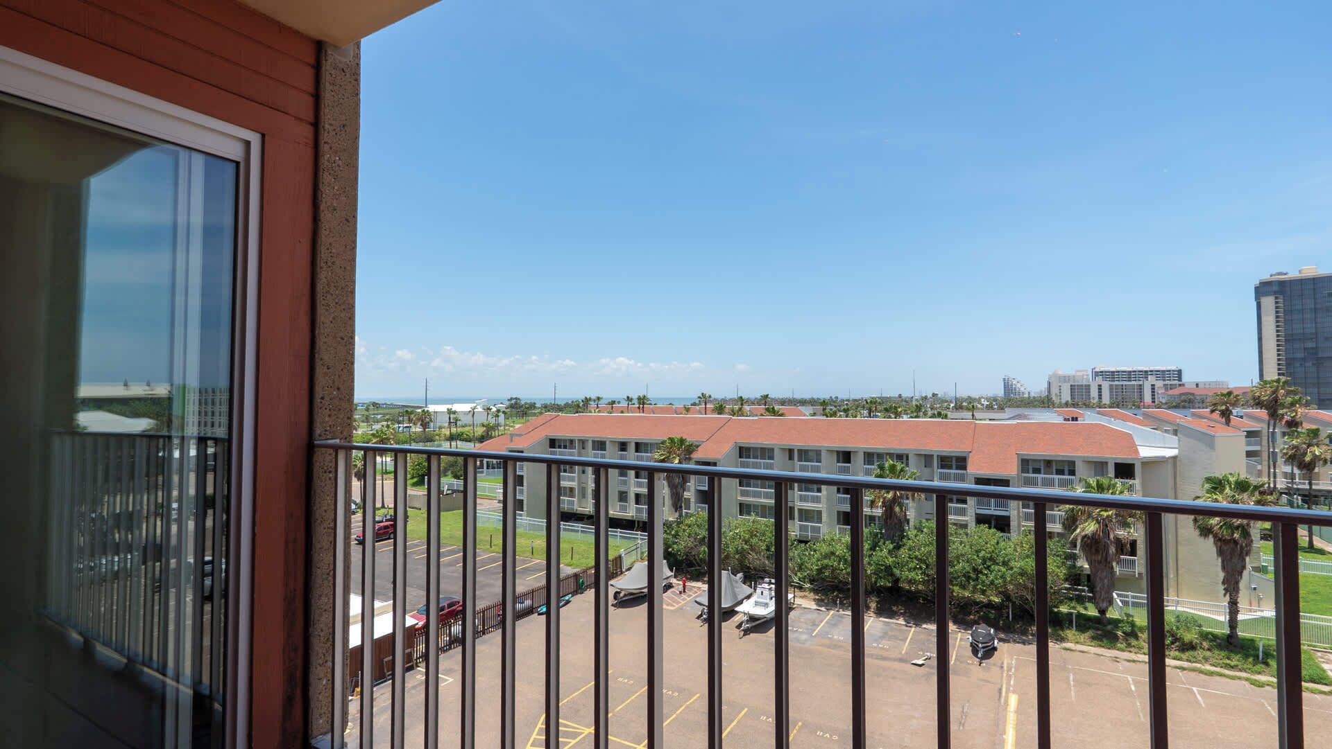 Un balcon donnant sur un parking, un immeuble avec un toit rouge et un ciel bleu dégagé.