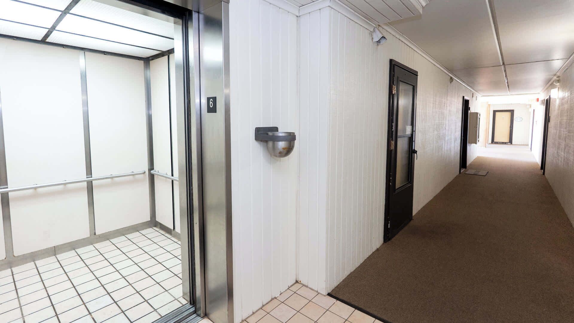 A hallway with a tiled floor, a door with a number plate, and a wall-mounted light fixture.
