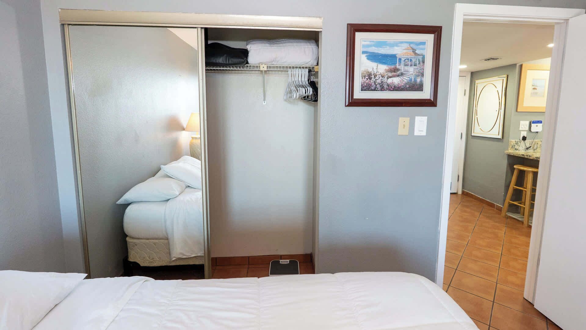 Een hotelkamer met een bed, een kledingkast met daarin hangende kleding, een schilderij aan de muur, en een zichtbare badkamer door een geopende deur.