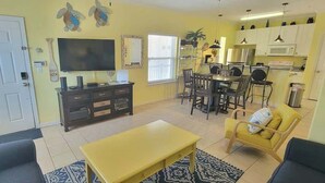 A living room with a yellow sofa, a coffee table, a TV, and a dining area with chairs and a table.