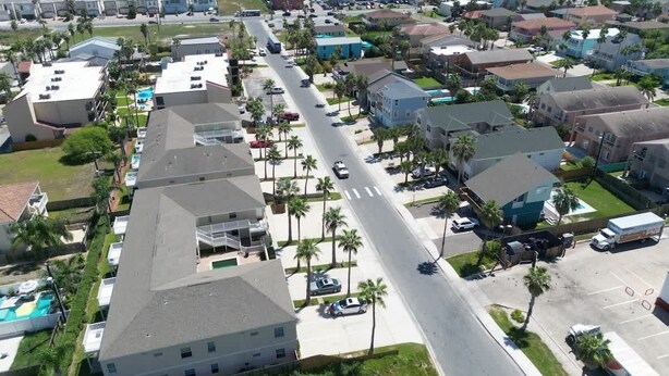 An aerial view of a coastal residential area with houses, a main road, and a beach in the background.
