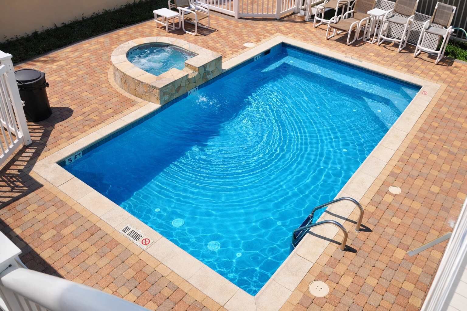 A rectangular swimming pool with a hot tub, surrounded by a tiled deck and white lounge chairs.