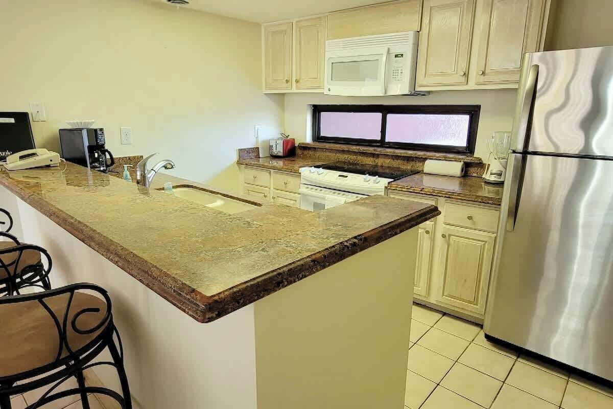 Una cocina con encimera de granito, armarios blancos, refrigerador de acero inoxidable, microondas, estufa y pileta.