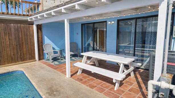 Un espace aménagé au bord de la piscine, avec une terrasse en bois, une table de pique-nique blanche et deux chaises Adirondack.
