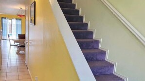 Un escalier avec une rampe blanche et des marches recouvertes d’une moquette violette, menant à l’étage supérieur.