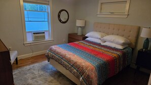 A bedroom with a bed, a nightstand, a lamp, a window with blinds, and an air conditioning unit. - Modern Condo #2! (Ellsworth)