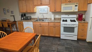 A kitchen with wooden cabinets, a microwave, a stove, a sink, and a dining area with a table and chairs. - Modern Condo #2! (Ellsworth)