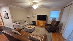 A living room with a brown leather sofa, a grey sofa, a wooden cabinet with a TV, a ceiling fan, and a window with curtains. - Modern Condo #2! (Ellsworth)