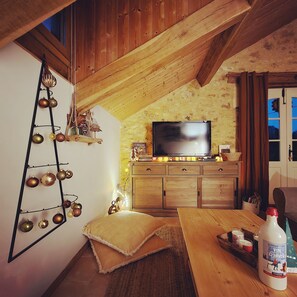 A cozy room with a wooden ceiling, a television on a wooden cabinet, a ladder with ornaments, and a bottle of wine on the table.