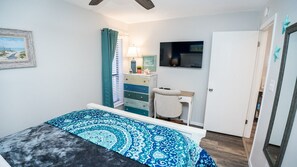 Ein Schlafzimmer mit einem Bett, einem Schminktisch, einem Schreibtisch mit Stuhl, einem Fernseher und einer Tür.