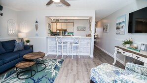 Ein modernes Wohnzimmer mit einem blauen Sofa, einem Holztisch und einem gemusterten Teppich. Es gibt eine Küchenzeile mit einer Bar und Hockern, sowie einen Essbereich mit einem weißen Tisch und Stühlen.