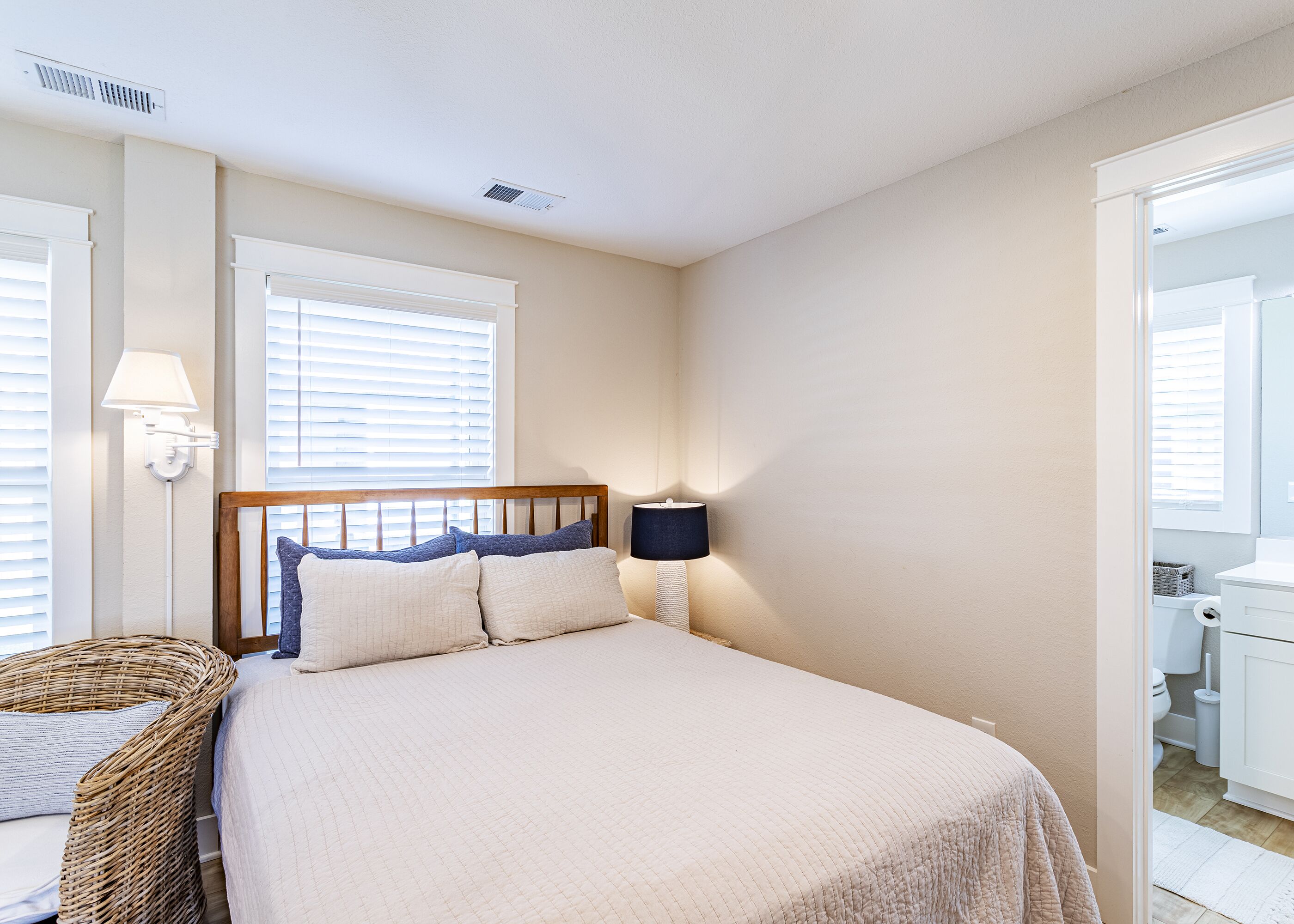 A neatly arranged bedroom with a bed, bedside lamps, a wicker basket, and a view into the bathroom.