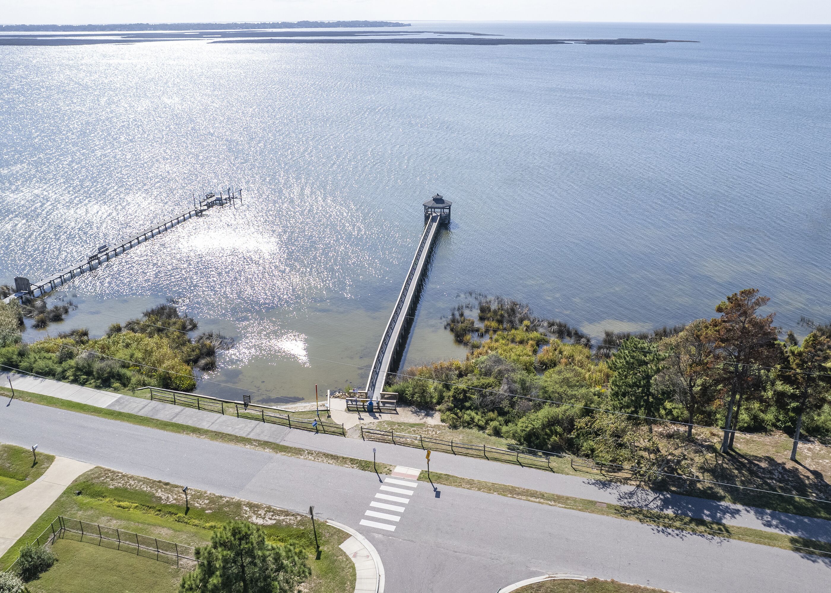 A waterfront promenade with a long pier extending into the water, surrounded by greenery and a clear sky.