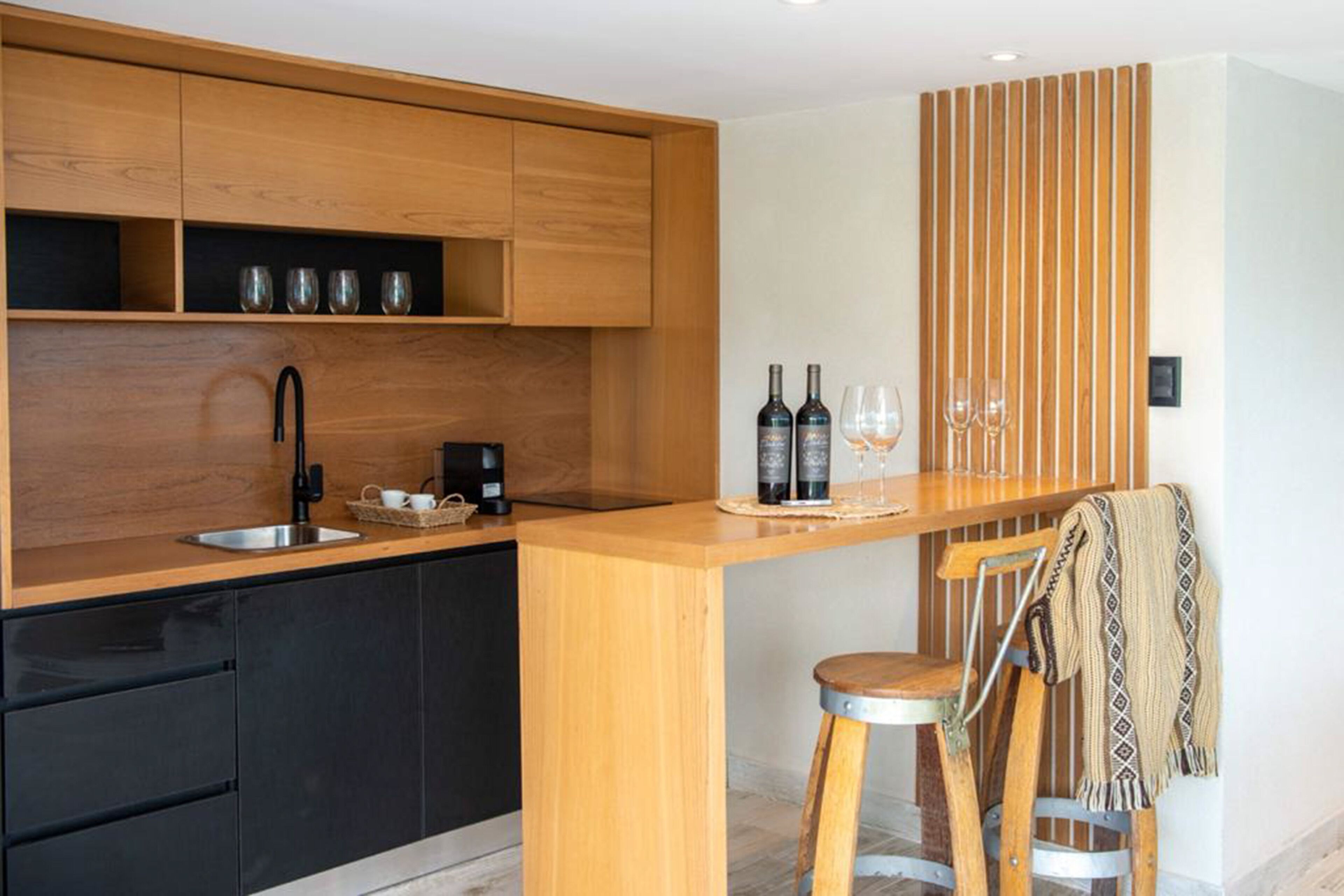 A modern kitchen with wooden cabinets, a black countertop, a sink, and a bar area with stools and wine bottles.