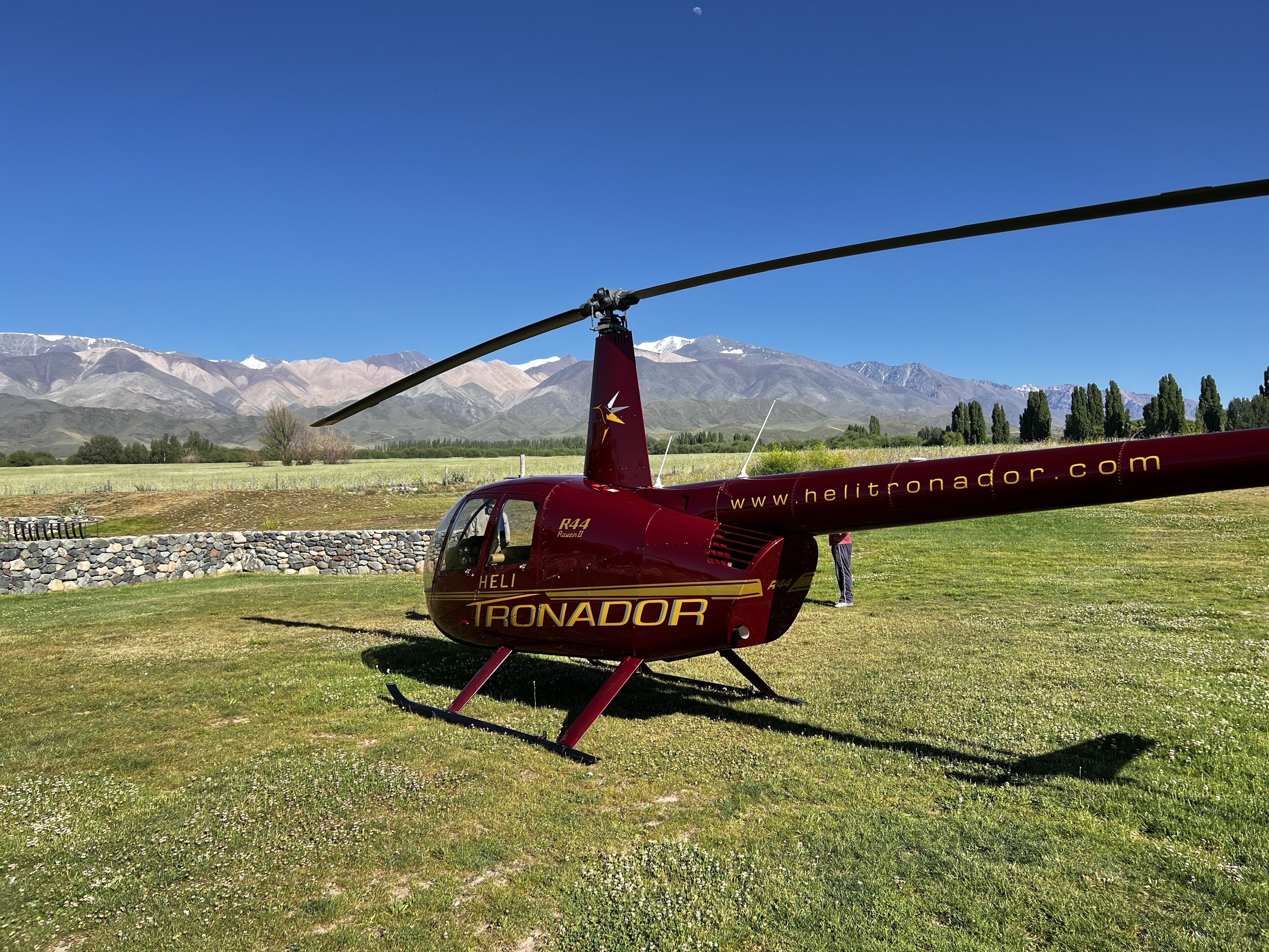 Un hélicoptère rouge, portant les inscriptions « Helitron.com » et « Heli Ironador » sur sa carlingue, est stationné sur un champ herbeux, avec des montagnes en arrière-plan.