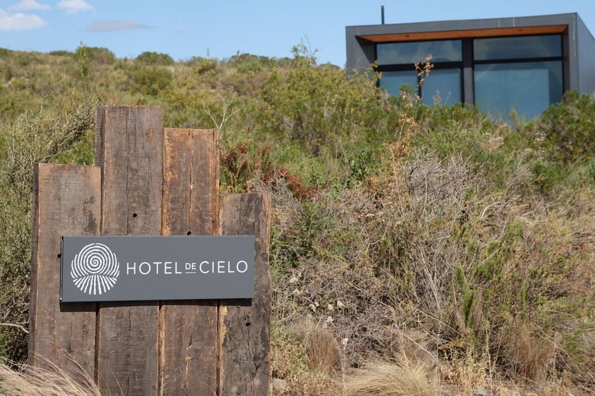 Ein hölzernes Schild mit der Aufschrift "HOTEL DE CIELO" und ein modernes Gebäude im Hintergrund.