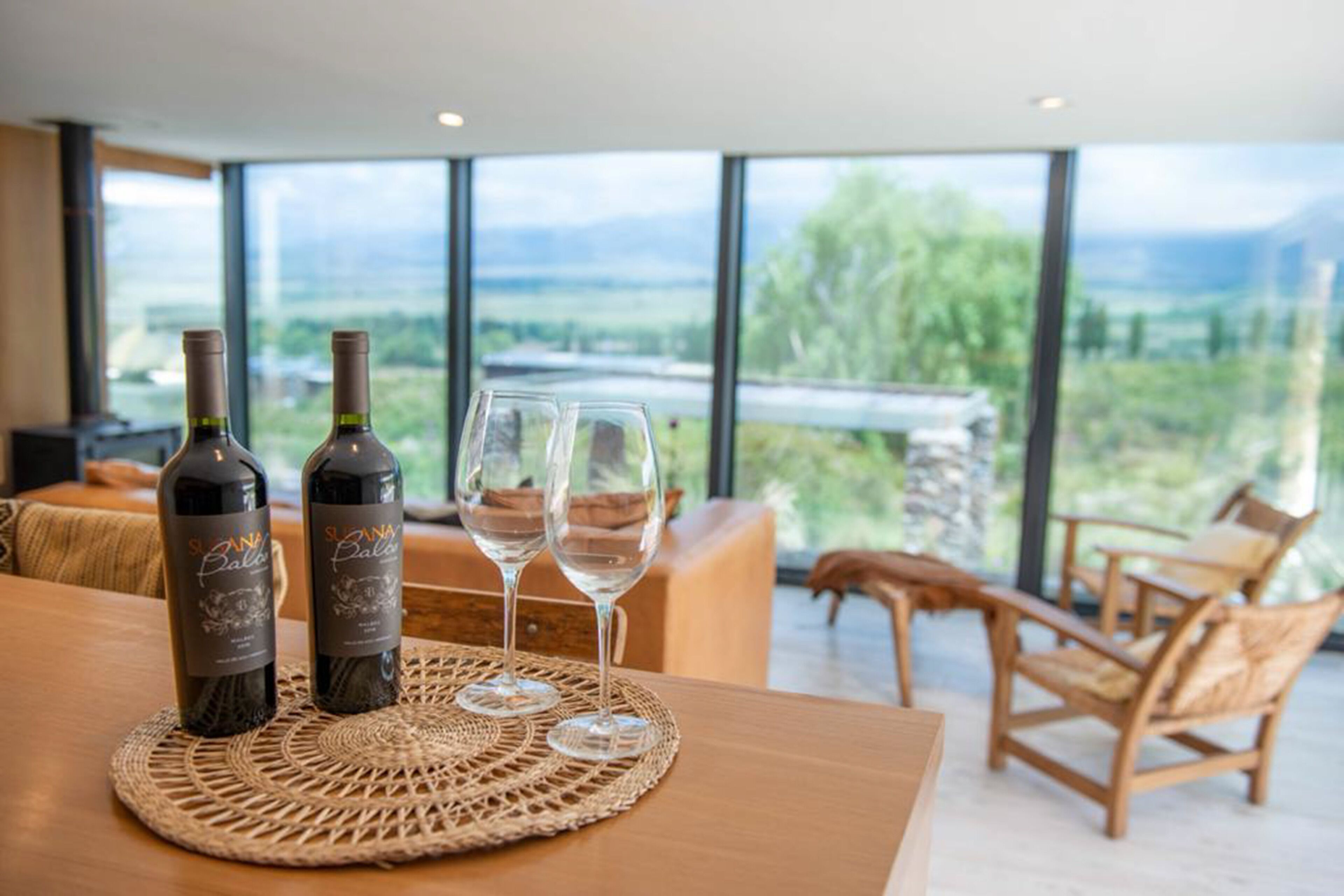 A table with two wine bottles and two glasses on a woven placemat, overlooking a scenic view through large windows.