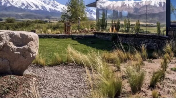 Une maison moderne en verre, dotée de fondations en pierre, entourée d’un paysage naturel avec des montagnes enneigées en arrière-plan.