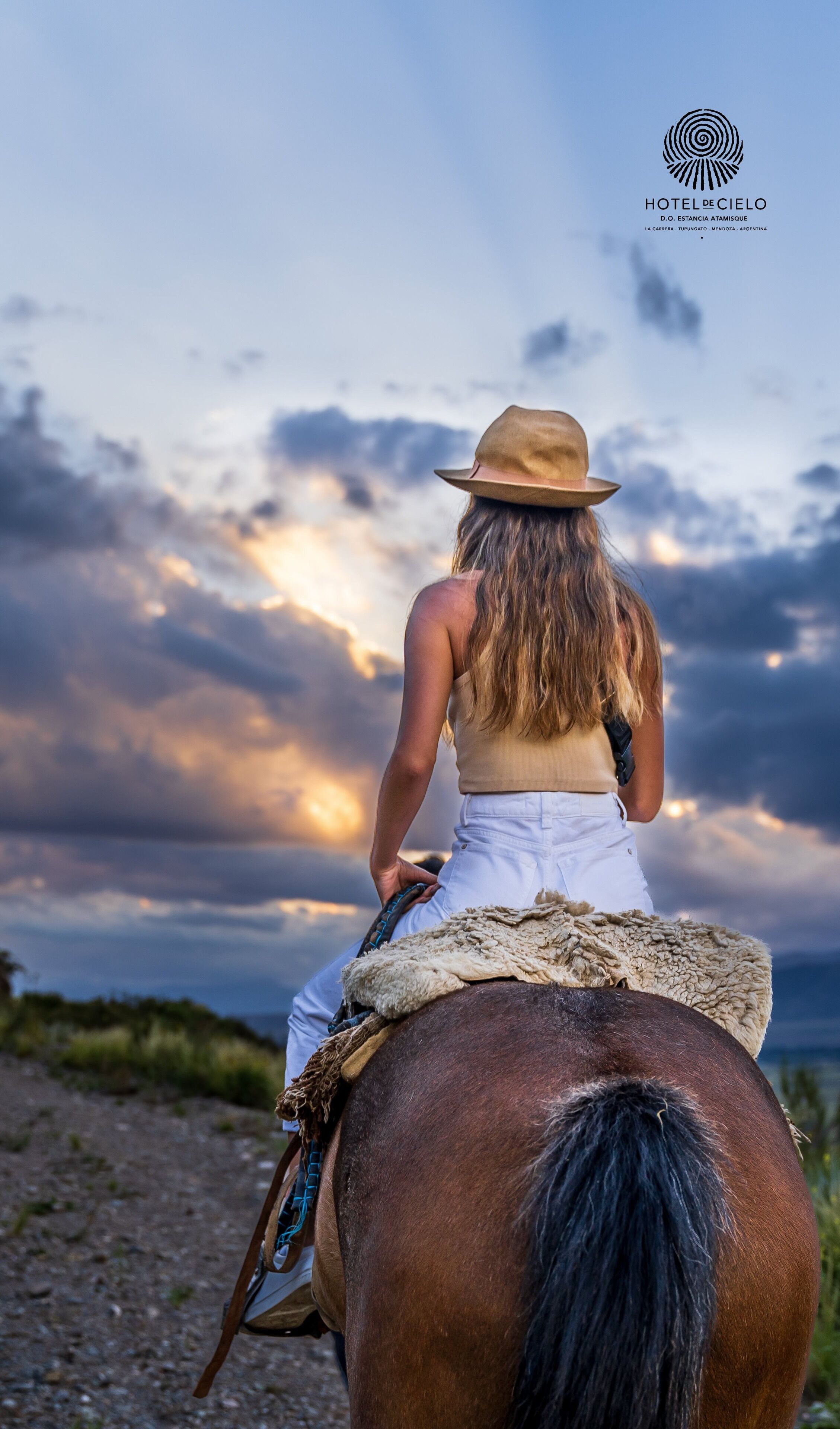 A person riding a horse with a saddle, wearing a hat, and a sleeveless top, with a logo of HOTEL DE CIELO in the upper right corner.