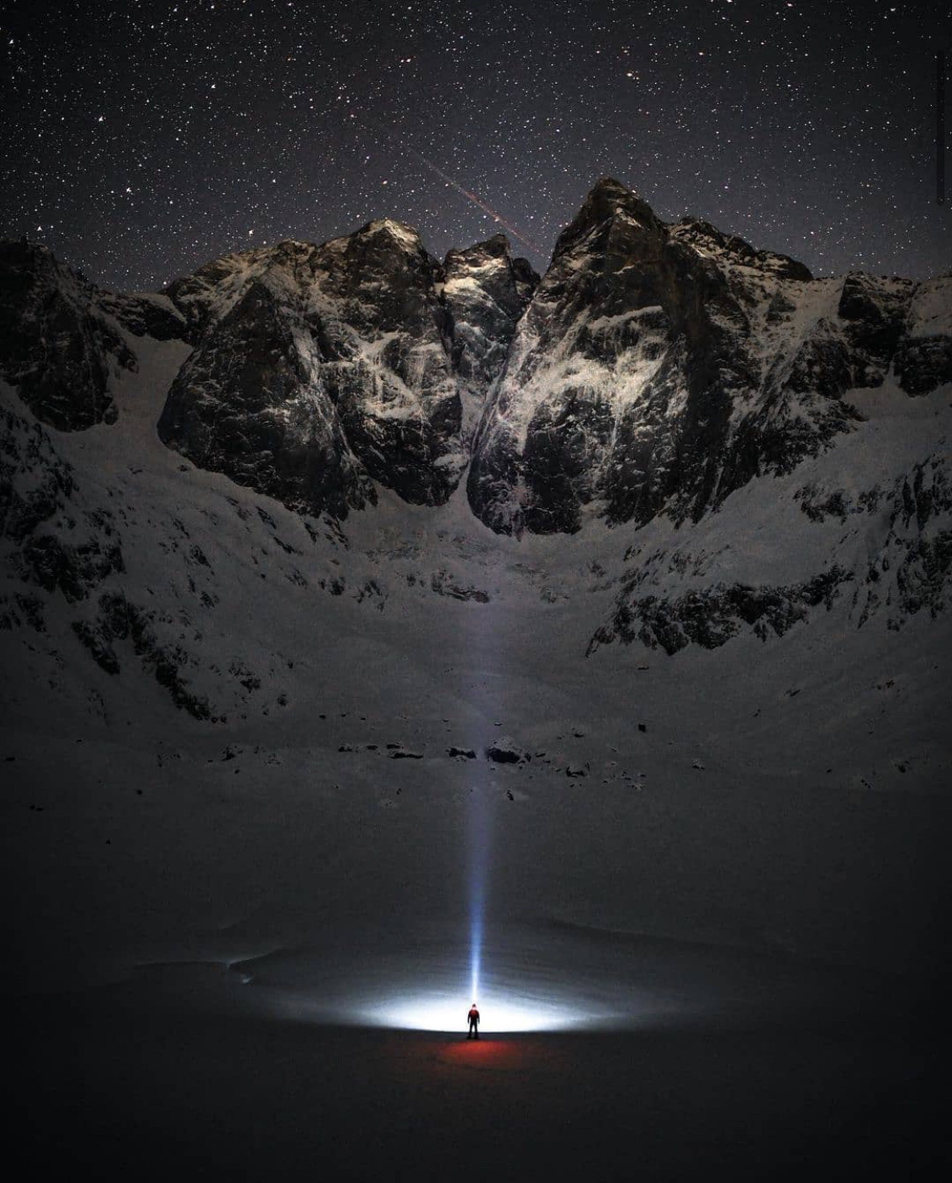 Eine Person steht in einem schneebedeckten Tal mit einem Bergkamm im Hintergrund und einem sternenübersäten Nachthimmel.