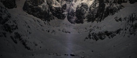 Eine Person steht in einem schneebedeckten Tal mit einem Bergkamm im Hintergrund und einem sternenübersäten Nachthimmel.