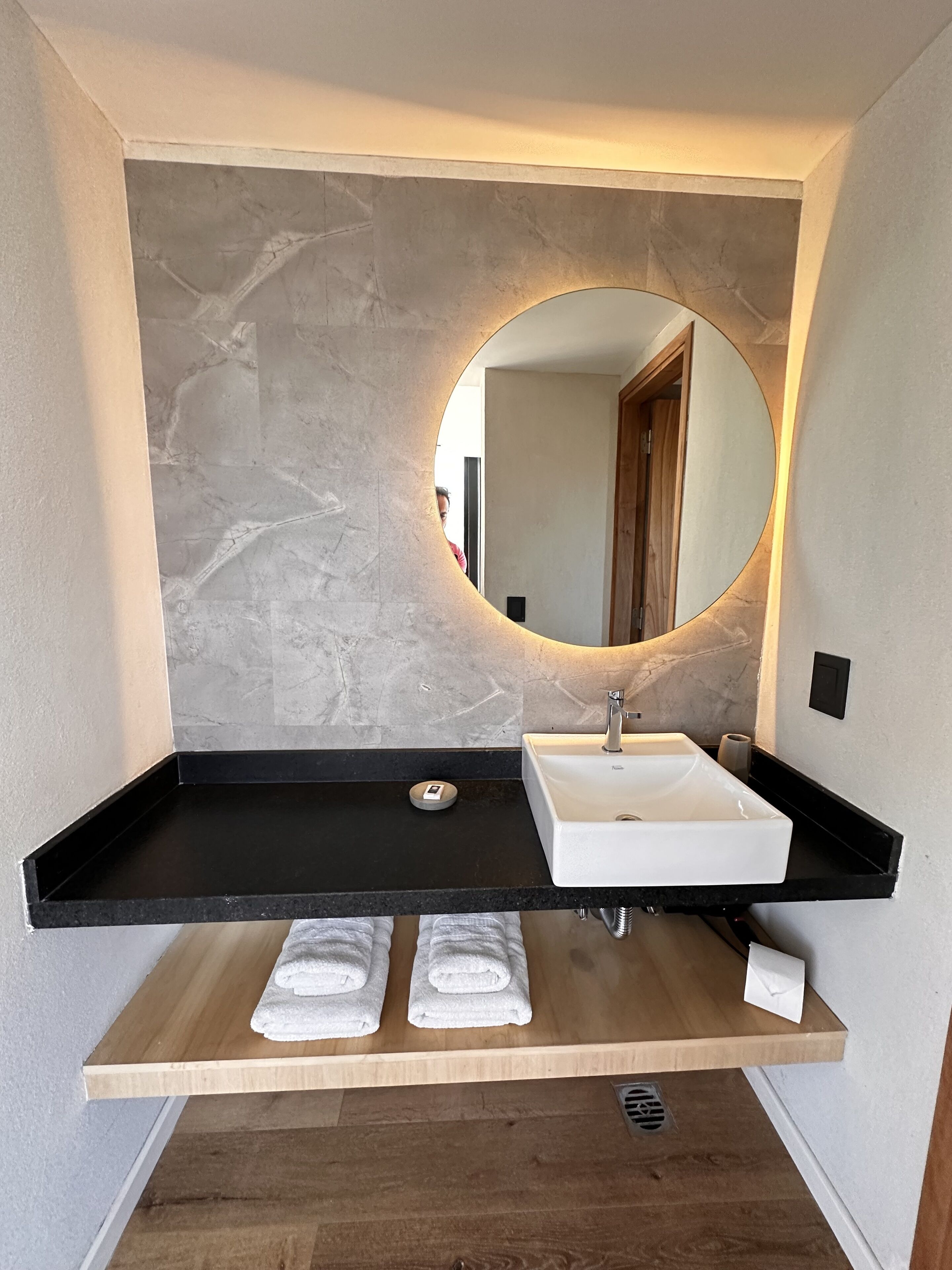 A modern bathroom with a marble wall, a black countertop, a white sink, and a wooden shelf with towels.