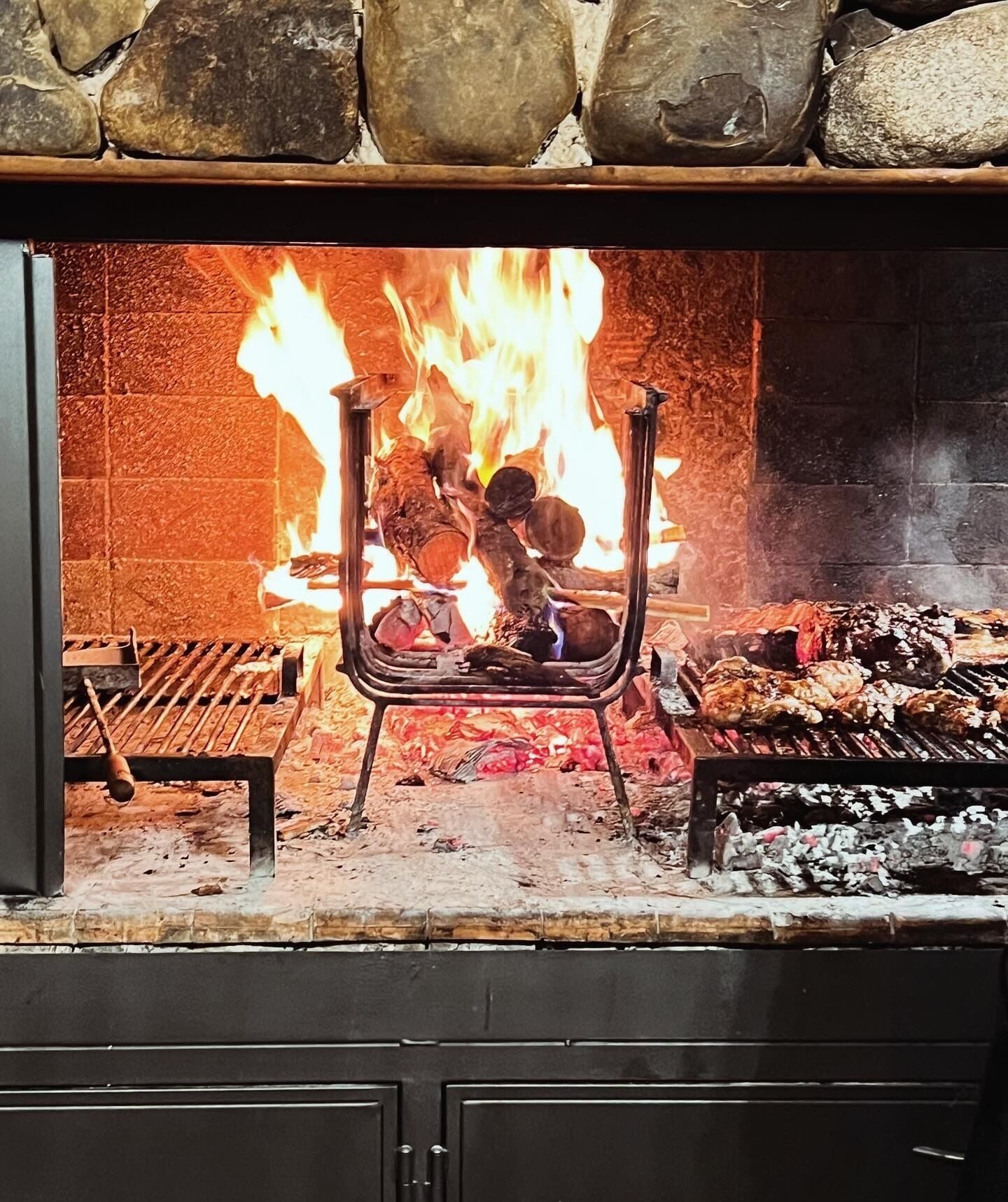 A fireplace with a roaring fire, a metal grill with food, and a stone wall.