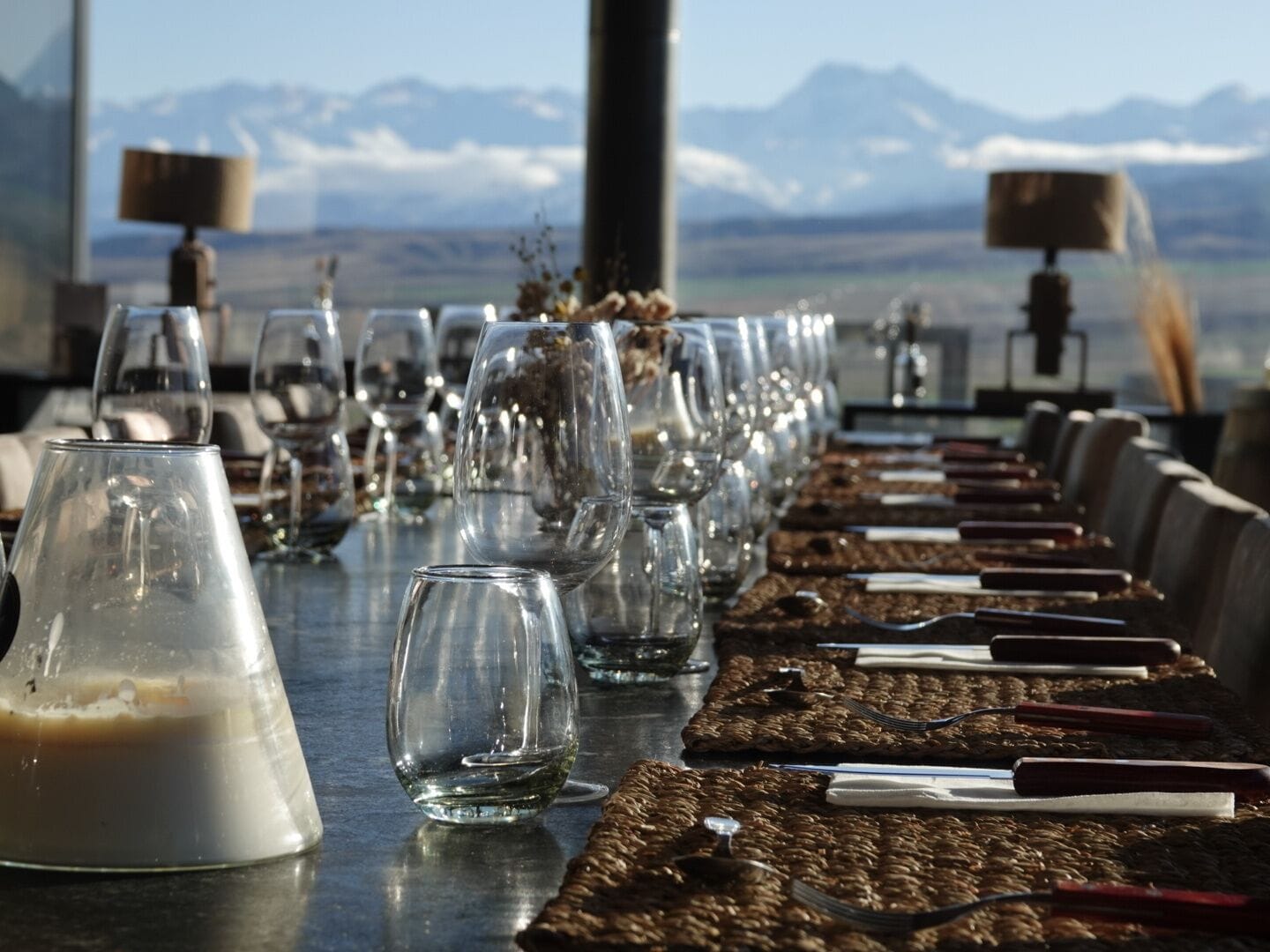 Una larga mesa servida con copas de vino, una jarra de leche y cubiertos, con una vista de montañas al fondo.