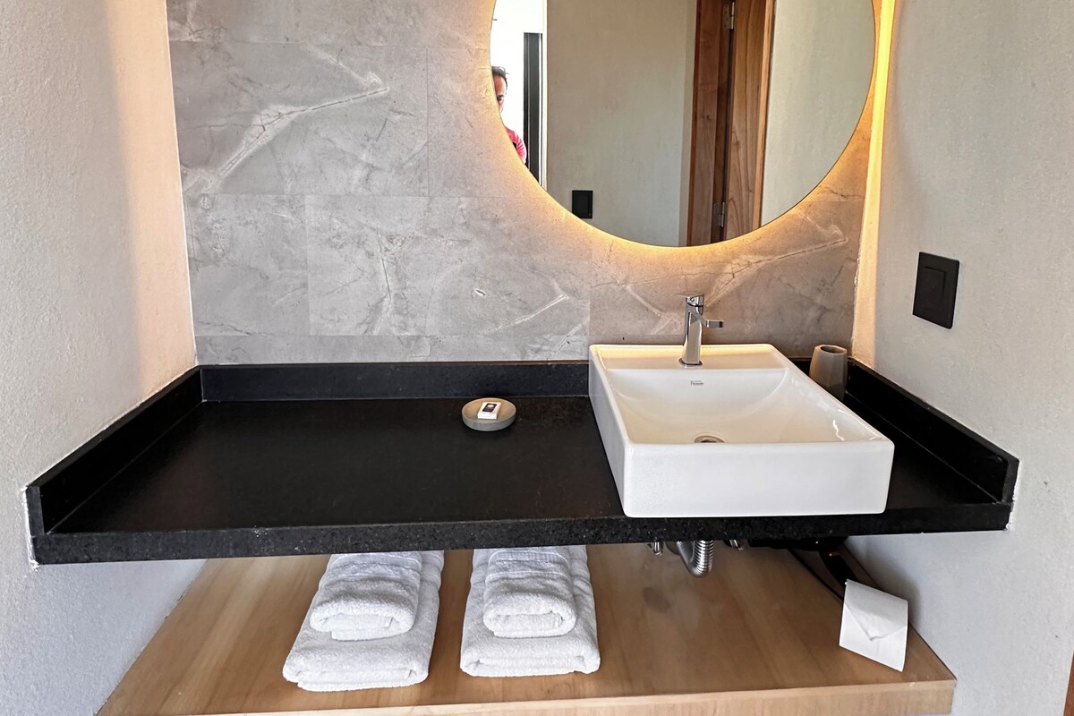 Un baño moderno con pared de mármol, encimera negra, lavamanos blanco y estante de madera con toallas.