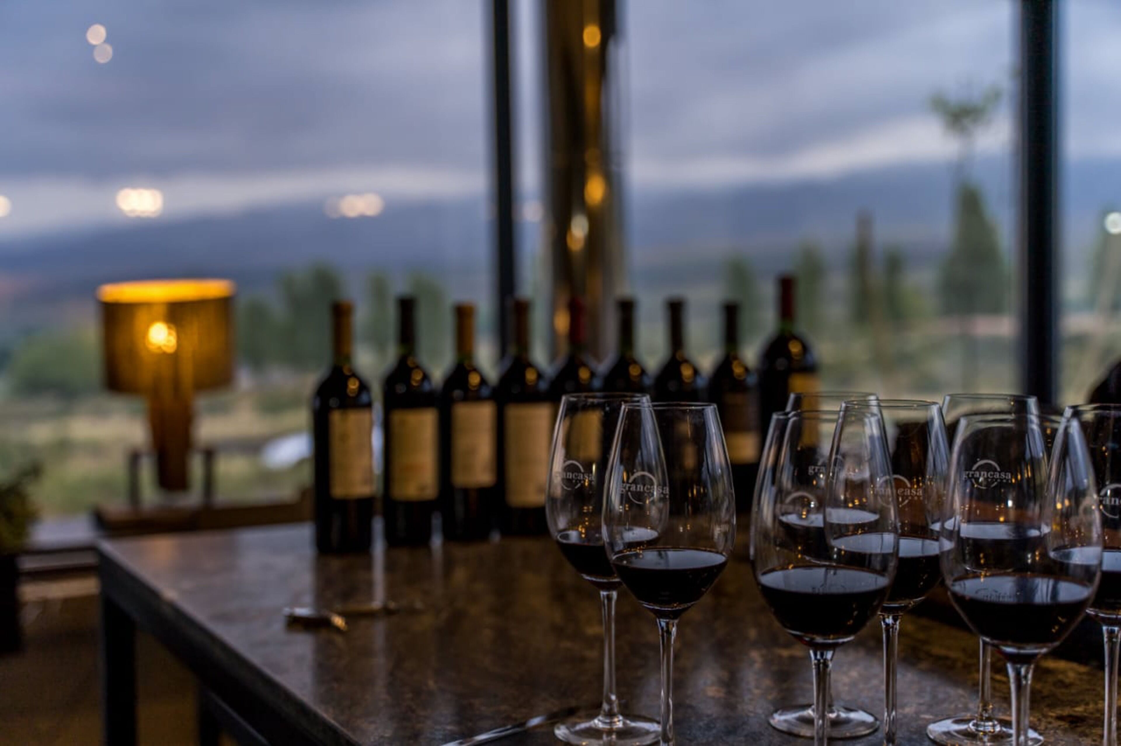 Un bar avec des bouteilles de vin et des verres, une lampe et une vue sur l’extérieur.