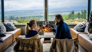 Une pièce chaleureuse avec une grande fenêtre donnant sur un paysage magnifique, une cheminée et une table où deux personnes trinquent avec des verres de vin.