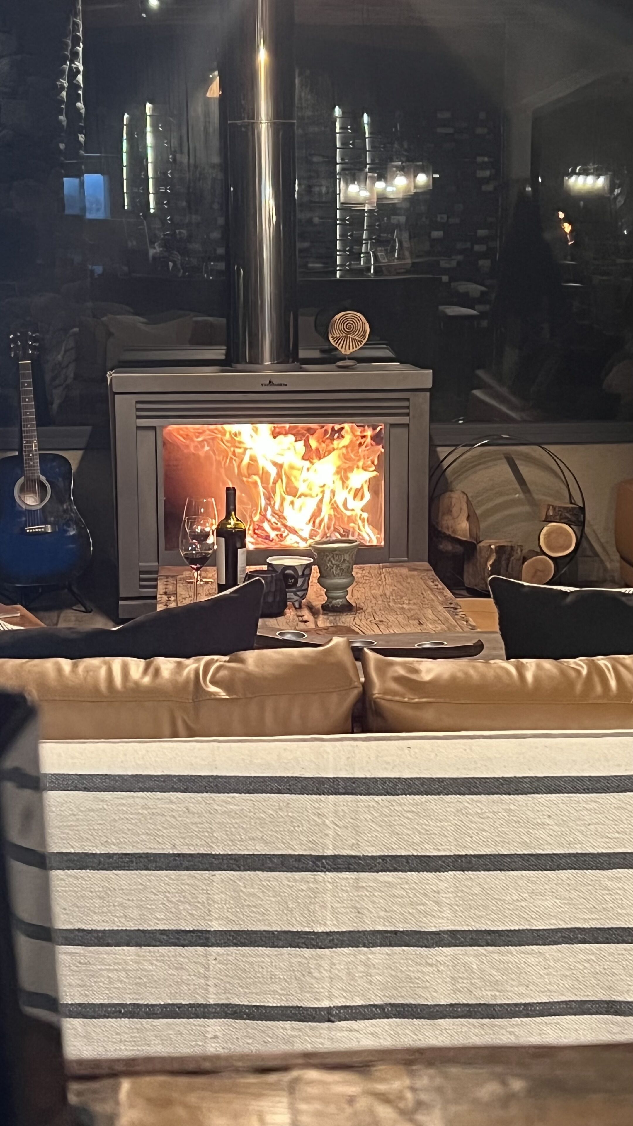 Un salon chaleureux avec un poêle à bois, une guitare et une table sur laquelle se trouvent une bouteille et un verre.