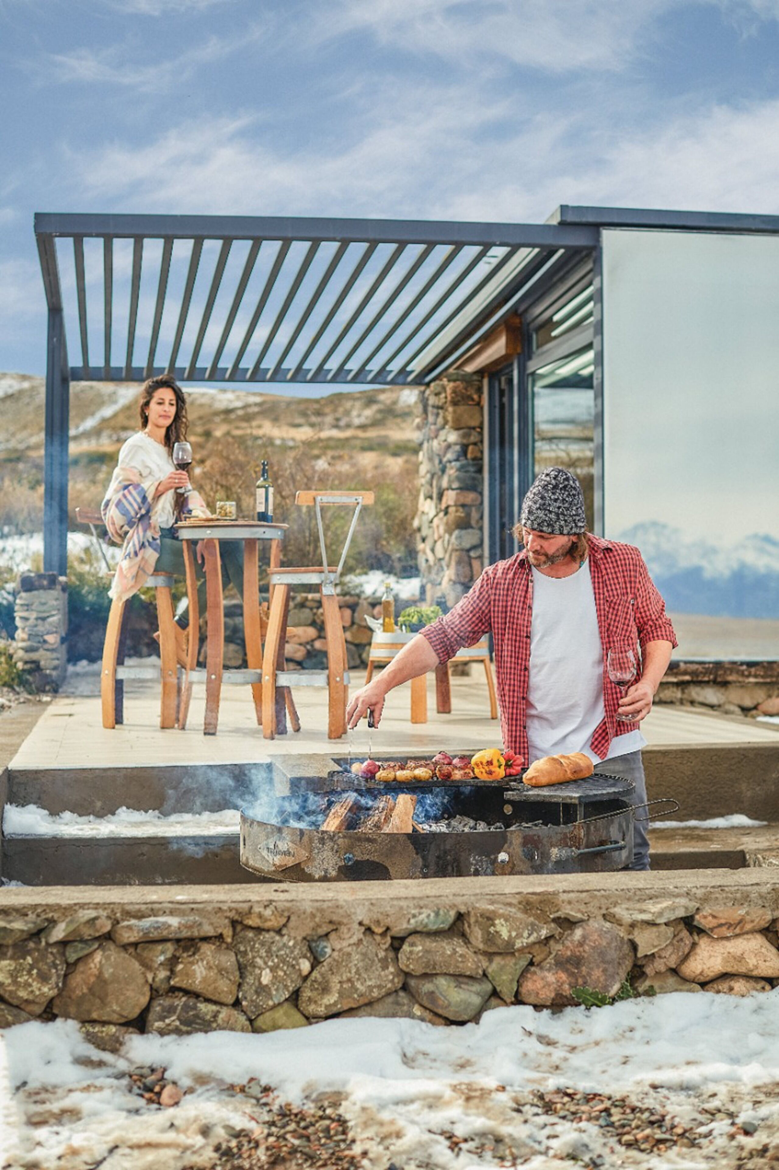 Een man barbecuet buiten, terwijl een vrouw op de achtergrond aan een tafel zit.
