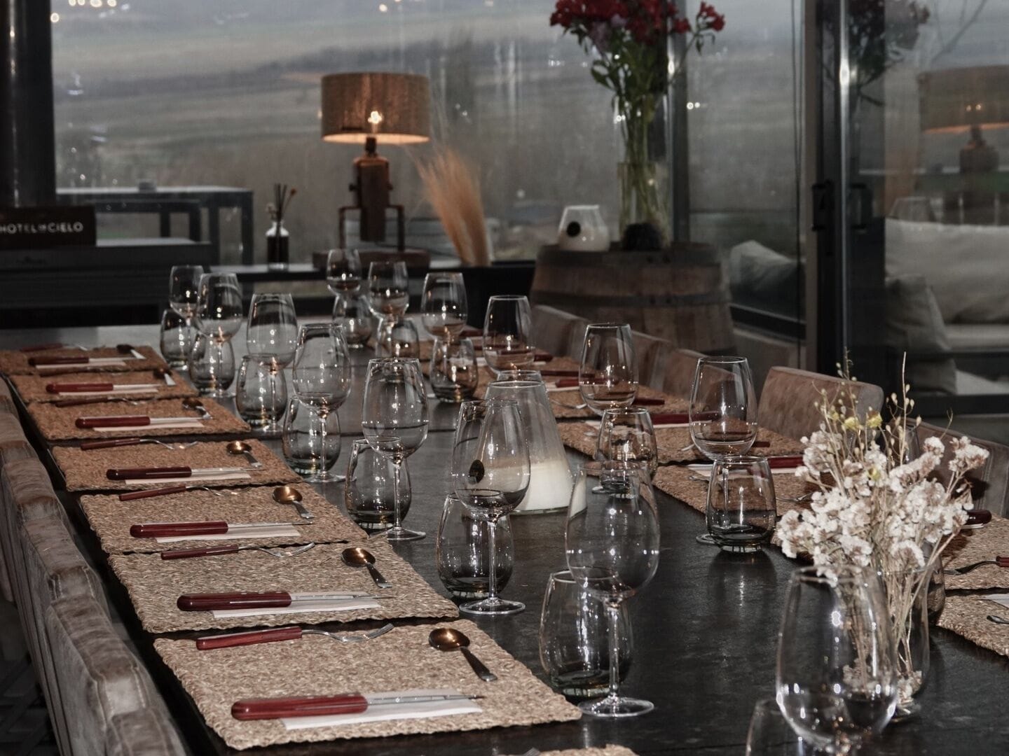 Una larga mesa de comedor dispuesta con múltiples copas de vino, cubiertos y vajilla, con vistas a la ciudad a través de grandes ventanales.