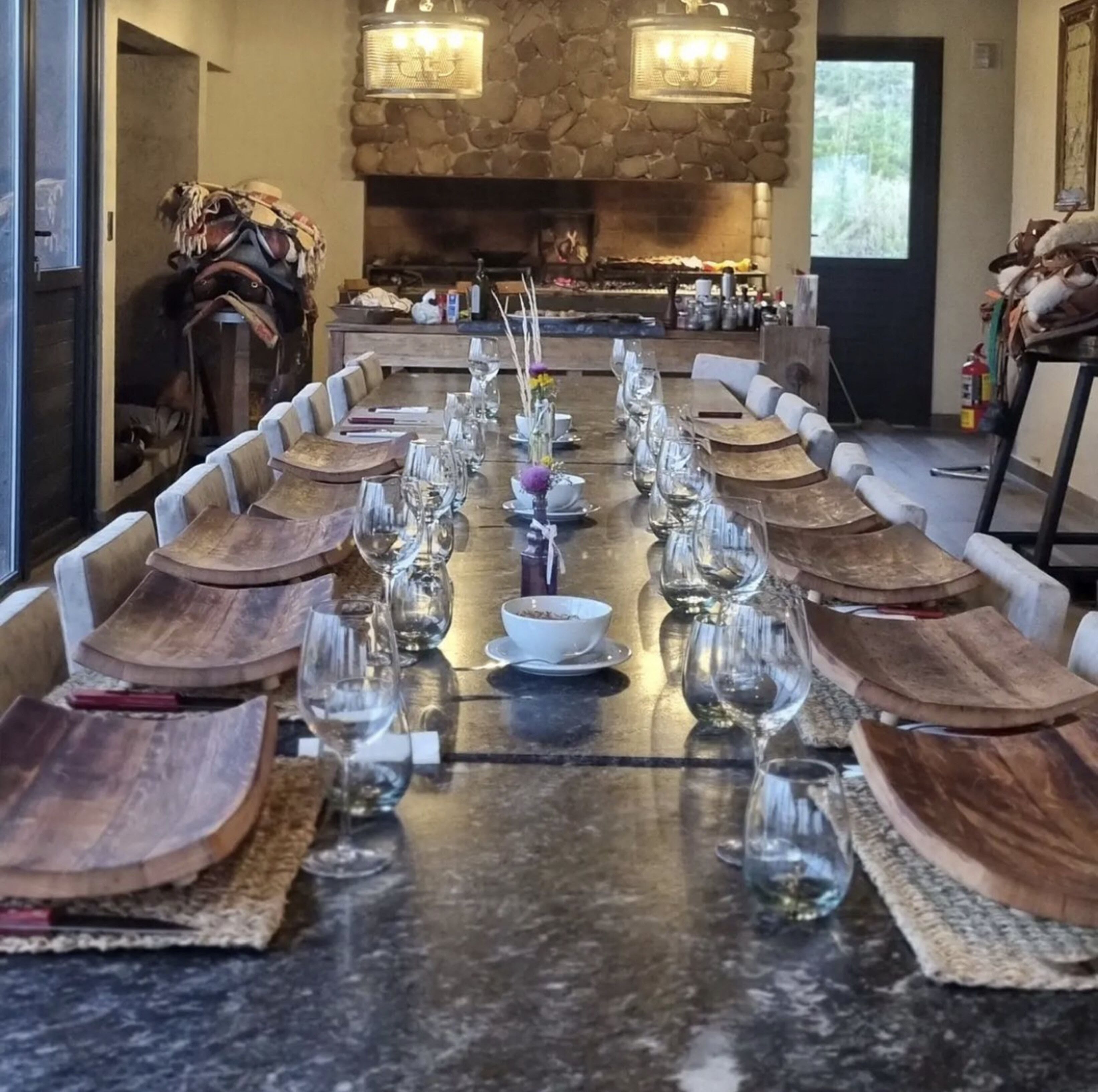 A long dining table with wooden cutlery and wine glasses, set in a room with stone walls and a fireplace.